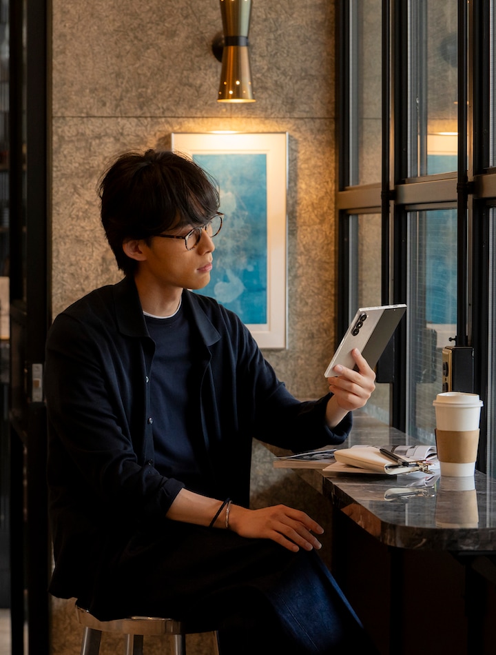 カフェの店内で座っている男性が、折りたたみスマホGalaxy Z Fold6（ギャラクシーZフォールド6）を片手に持ち、開いた画面を見ている様子。