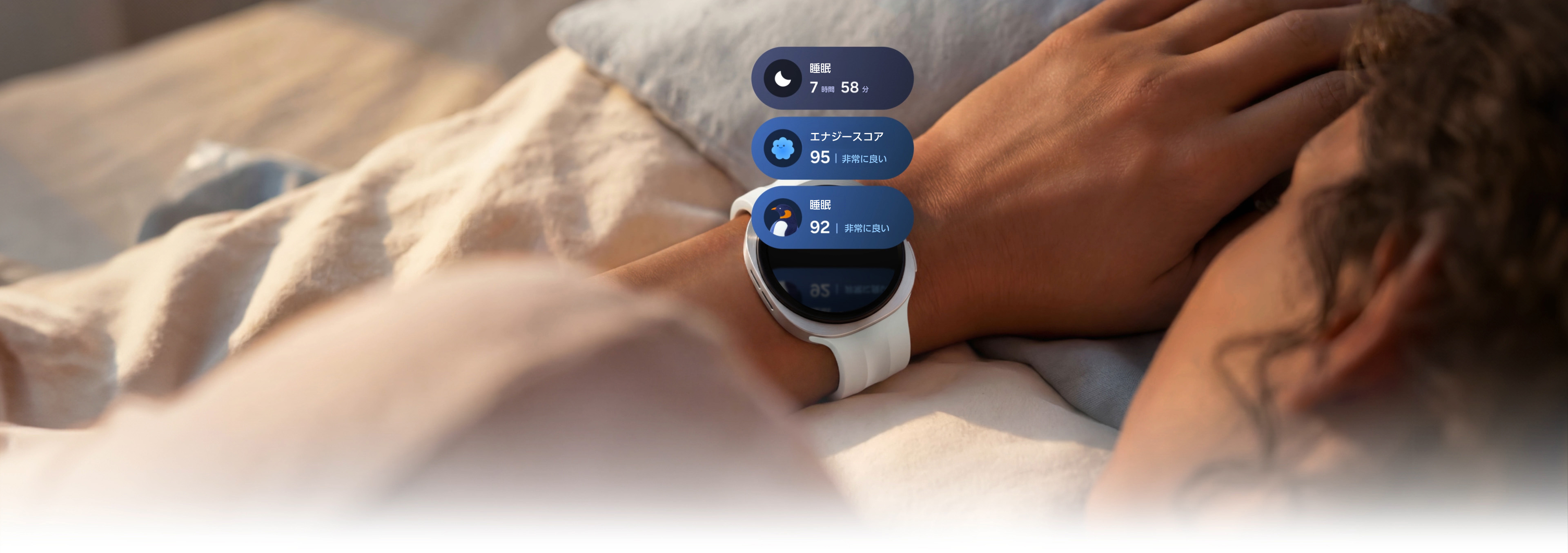 Galaxy Watch9（シルバー）にSport Band（クリーム）を手首に着けた画像。ウォッチのディスプレイ上部に3つの睡眠トラッキングタイルが表示されており、睡眠時間7時間58分のタイル、95という非常に良いエナジースコアのタイル、そしてペンギンのアイコンと92という非常に良い睡眠スコアが表示された睡眠コーチングタイルが含まれる。