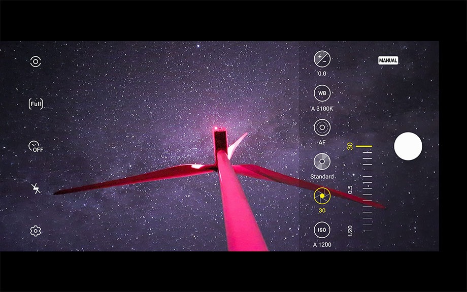A bright red wind power generator stretching up into the sky against some Milky Way with on-screen camera controls demonstrating how shutter speed works.