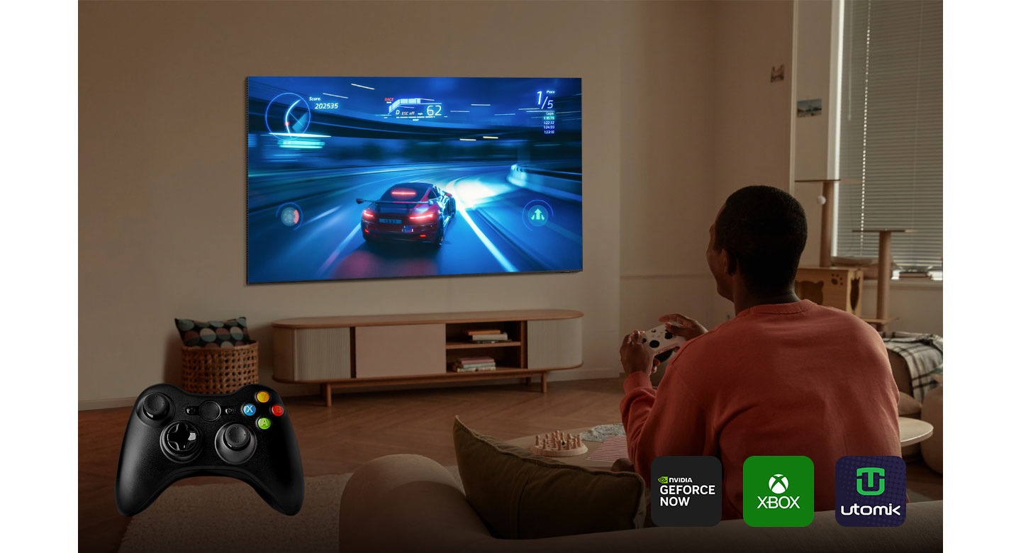 Мужчина увлечен игрой на экране своего телевизора Samsung Smart TV. В левом нижнем углу экрана на видном месте находится элегантный черный контроллер. На экране также представлены популярные игровые приложения, включая NVIDIA GeForce Now, Xbox и Utomik.