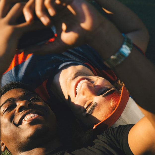 Two friends laying on the ground together in opposite directions, looking up at a smartphone one of the men is holding, smiling while the sun shines down upon them.