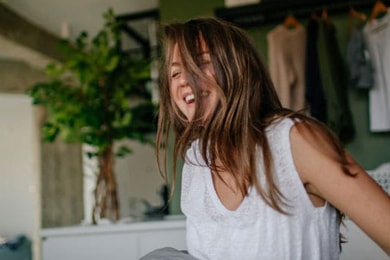 A woman sits up in bed in her room, laughing with her eyes closed as her hair covers half of her face. She is wearing a white sleeveless tank top.