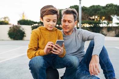 An outdoor scene of a young boy sitting on a basketball and looking at his phone with his father sat beside him