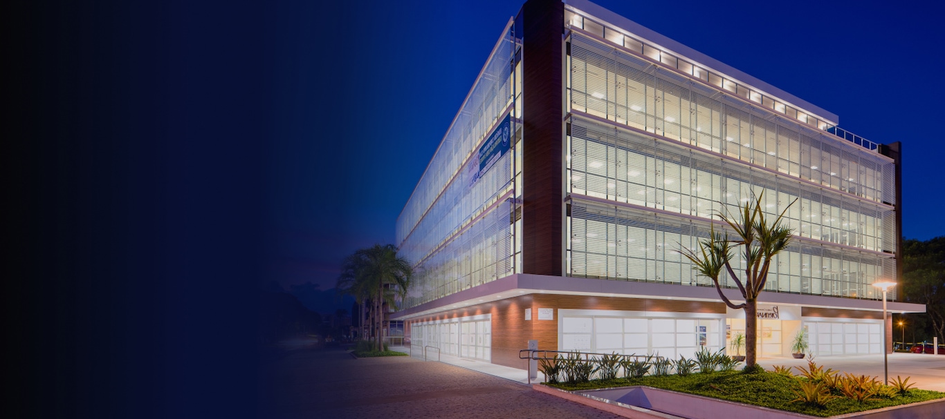A modern glass office building illuminated at night, with landscaping and palm trees in front, viewed from an angled perspective.