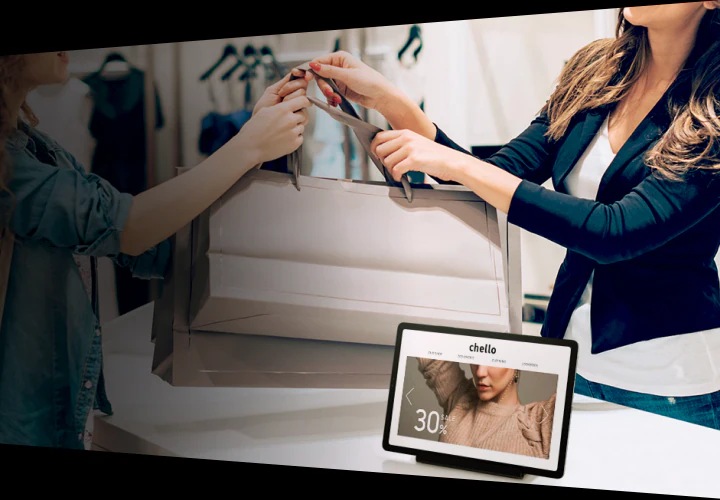 A customer at a clothing store is receiving her purchases from the store clerk. On the table between them is a Galaxy tablet device showing the store's online catalogue.