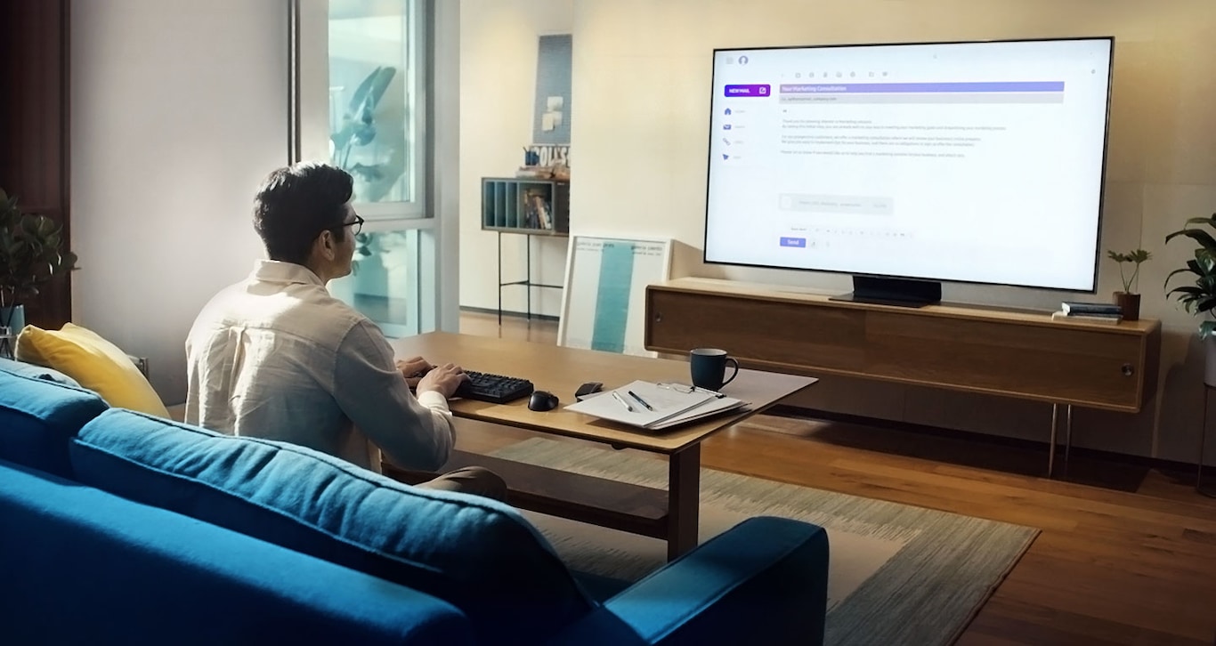Vyras sėdi ant sofos savo svetainėje ir spausdina klaviatūra, kuri prijungta prie „Samsung Smart TV“. Ekrane  nuotolinės prieigos pagalba  galime matyti, kas yra jo biuro kompiuteryje. Jis rašo el.laišką, kuris rodomas ekrane.