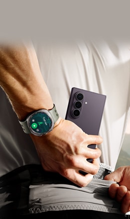 Un hombre guarda en su bolsillo un Galaxy Z Fold8 Ultra en color Violeta Sombra. En su muñeca está el Galaxy Watch Ultra2 en color Plata Titanio. El menú de entrenamiento Trail Run aparece en la pantalla del reloj.
