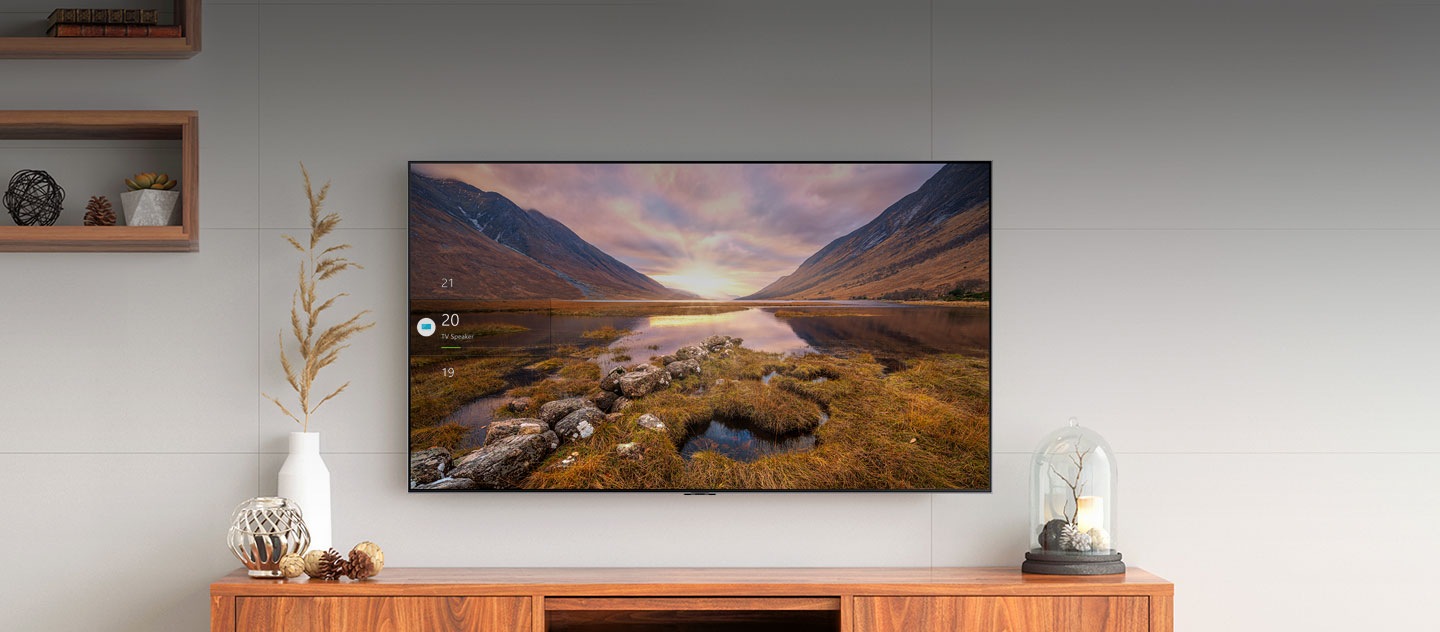 At the center of the living room, a large Samsung TV is hung up on the wall displaying a majestic view of nature. Below it lies a TV stand and various interior decorations.