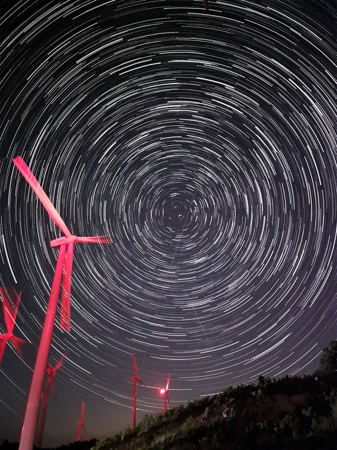 Long exposure shot of the stars in the night sky, forming a circular pattern that fill the sky. There are six red wind power generators in the foreground facing the left.