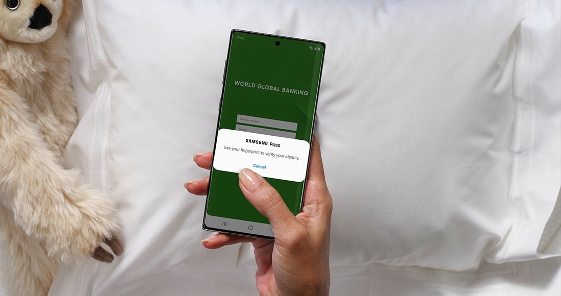 A hand holding Galaxy Note10 plus with the Samsung Pass interface onscreen. The person is using their phone in bed to access their bank account and Samsung Pass securely keeps their password so they can use biometric authentication to log in