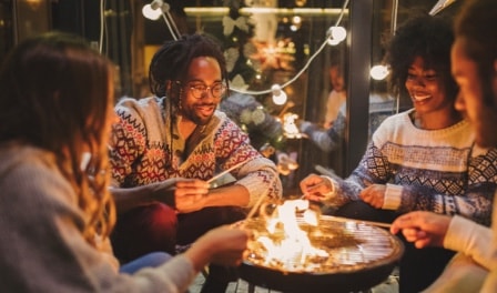 Un groupe de quatre adultes assis à l’extérieur fait griller des guimauves autour d’une table à feu.