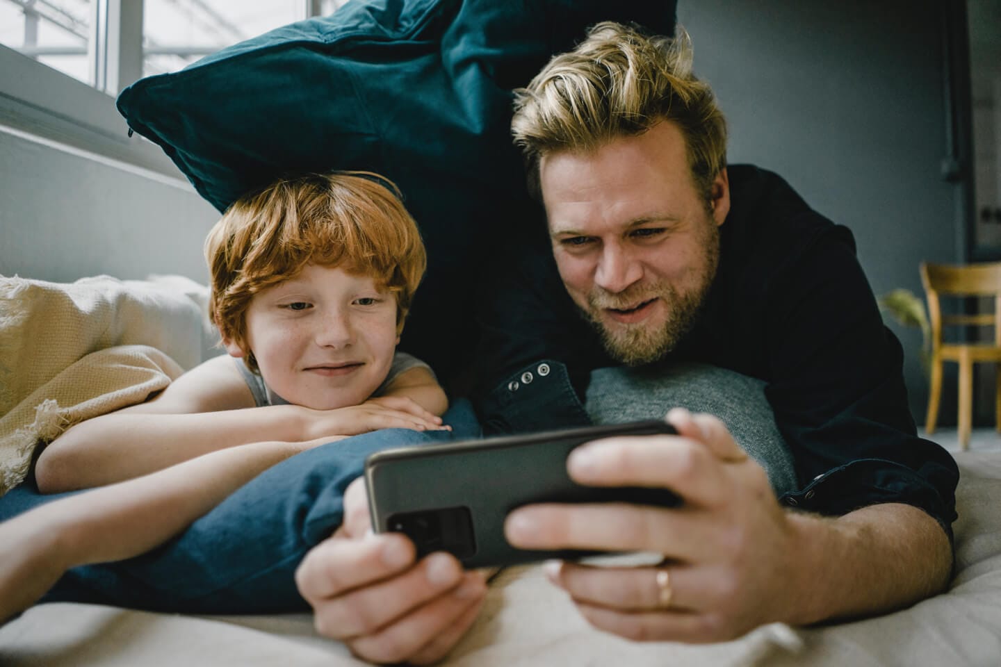 Un père et son fils allongés sur le ventre ; le père tient un smartphone gris foncé en mode paysage. Ils sourient tous les deux en regardant l’écran du téléphone comme s’ils admiraient un spectacle à la maison.