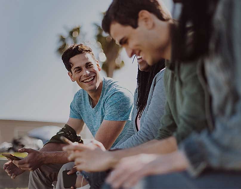 En gruppe unge mennesker kan ses sitte med smarttelefonene i hendene. Et av gruppens medlemmer smiler til de andre.