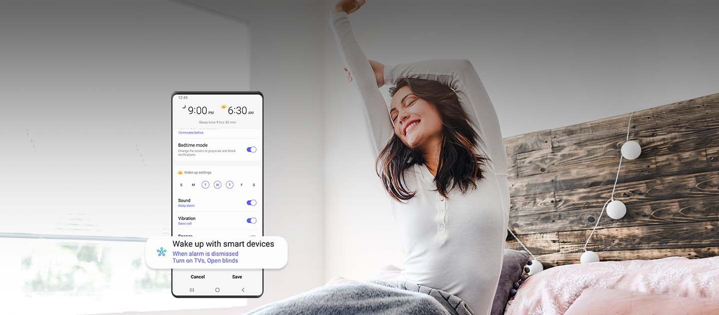 A woman waking up, stretching on the bed with a smile. On the foreground, a Galaxy smartphone GUI shows the duration of sleep and other enabled menus including, Bedtime mode, Sound, Vibration, etc. Towards the bottom of the GUI, a certain menu is enlarged. It reads, "Wake up with smart devices. When alarm is dismissed, turn on TVs, open blinds."