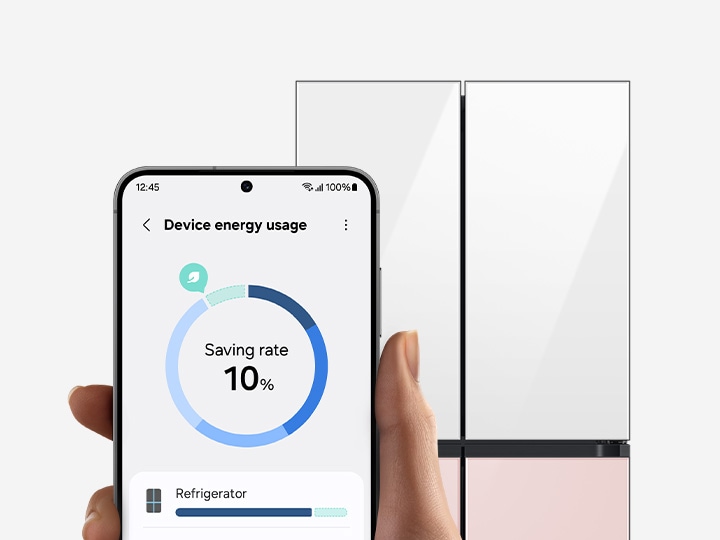 In front of a refrigerator, a hand holds a Galaxy smartphone displaying the AI energy mode SmartThings app UI.