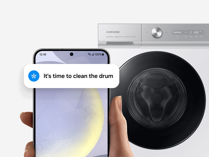 In front of a washing machine, a hand holds a Galaxy smartphone displaying the SmartThings app UI with drum clean notification.