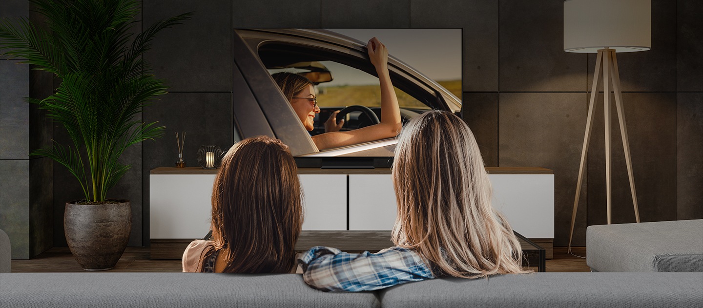 In the foreground, we see the back of the heads of two women - one with blonde hair and the other with red hair. They are watching a show on a large, living room TV in the background.
