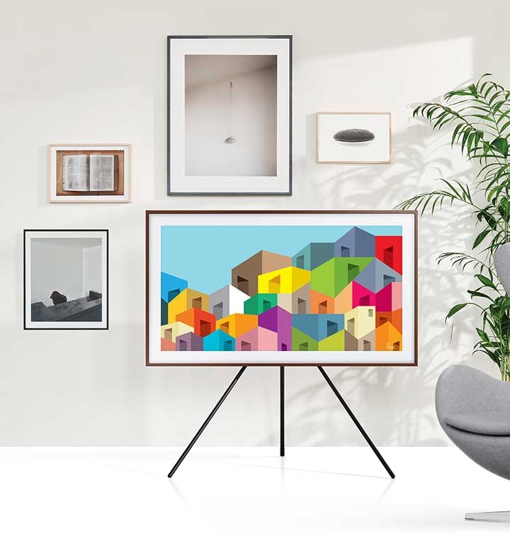 The Frame is mounted on a stand and is displaying a variety of artworks. The Frame is located next to pictures framed on the wall, which makes The Frame also look like a picture frame.