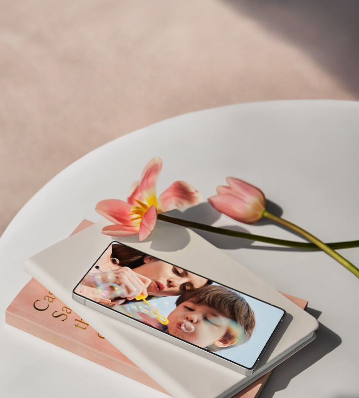 A Galaxy S25 Ultra lies on the table on top of a stack of books. The screen displays a photo showing a mother and child blowing bubbles together. Beside the books there are two pink tulips, one fully bloomed and the other still partially opening.