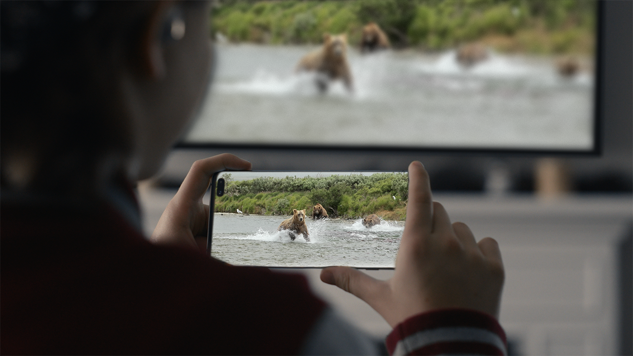 A young girl is holding a Galaxy Note10 in her both hands. And there is a video of bears in the water scene on her phone that is also on the TV in front of her.