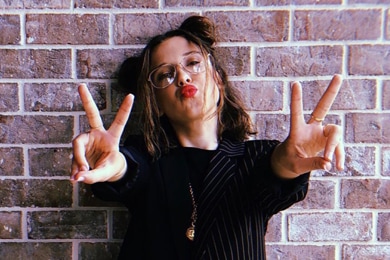 Millie Bobby Brown, de pie contra una pared de ladrillos con un blazer negro a rayas y pantalones rojos, y formando dos signos de paz con las manos mientras frunce los labios para la cámara.