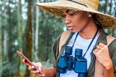 Una mujer con un sombrero de ala ancha y binoculares alrededor del cuello mira su teléfono mientras se apoya sobre un árbol en el bosque.