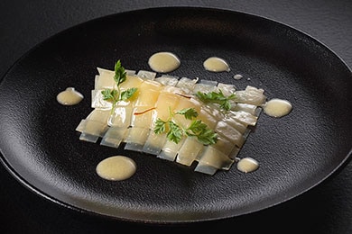 En el centro de un plato negro redondo, se superponen tiras de papa en capas sobre el bacalao que se trituró cuidadosamente. Para completar el elegante plato, se coloca salsa blanca sobre la preparación y alrededor en el plato como decoración, todo con un toque de perejil francés.