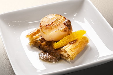En un plato cuadrado y blanco, se encuentra una porción de pissaladière coronada con una rodaja de naranja y una vieira. Un poco de pasta de aceitunas a la naranja completa el emplatado sencillo pero elegante.