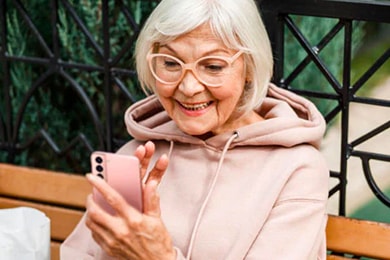 Una mujer mayor con un suéter rosa está sentada en un banco, con un teléfono Samsung Galaxy S21.