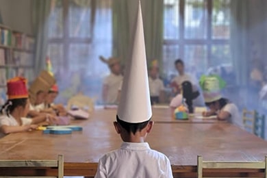 Una escena de la película de Sha Mo, Kids of Paradise, en la que un niño pequeño usa un gran sombrero cónico de punta en un salón de clases con otros niños.