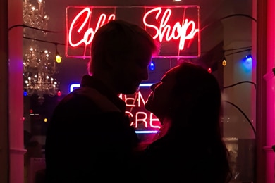 Fotografía de siluetas de una pareja parada en frente de una luz de neón de una cafetería, la escena es oscura pero se puede resaltar el contorno