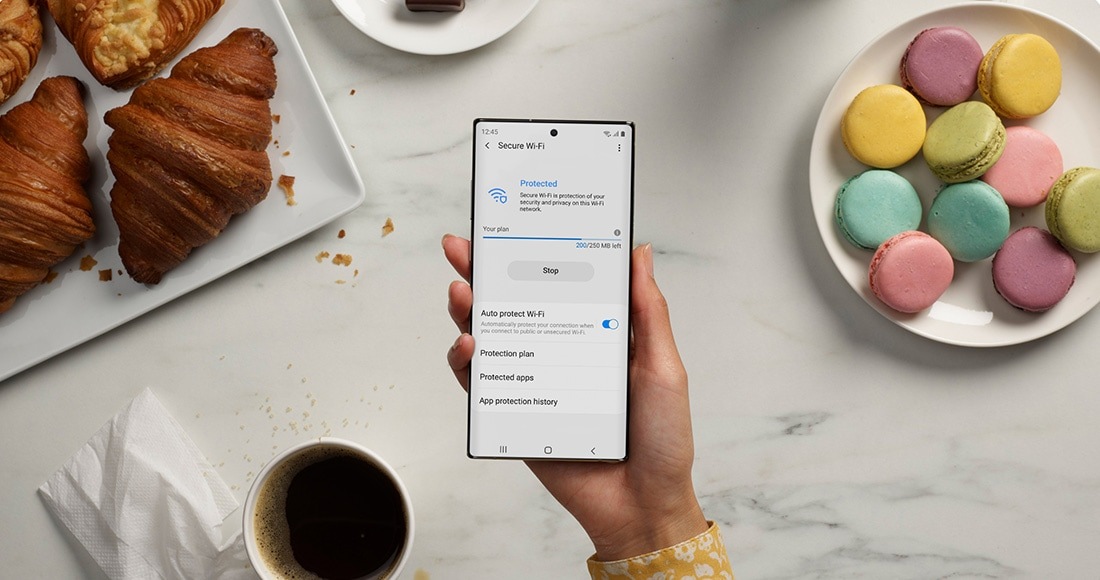 Ruka koja drži Galaxy Note10 plus sa Bezbednim Wi-Fi interfejsom na ekranu. U pozadini je tanjir sa kroasanima, tanjir makaronasa i šoljica kafe, koji predstavljaju lako obezbeđenje koje Knox pruža čak i kada je telefon povezan na javnu Wi-Fi vezu u kafiću