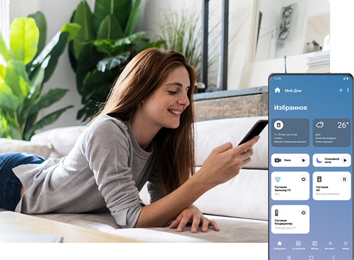 Женщина лежит на диване, улыбается и смотрит на свой смартфон. Справа на переднем фоне показан экран Galaxy с интерфейсом приложения SmartThings, где подключены различные умные устройства, виден их статус и другие операции, которые можно настраивать, включая «доброе утро» и «спокойной ночи».