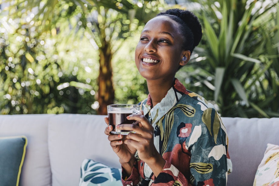 A woman sits on an outdoor couch with a beverage in her hand. Plants and trees lit by the sun are behind her.