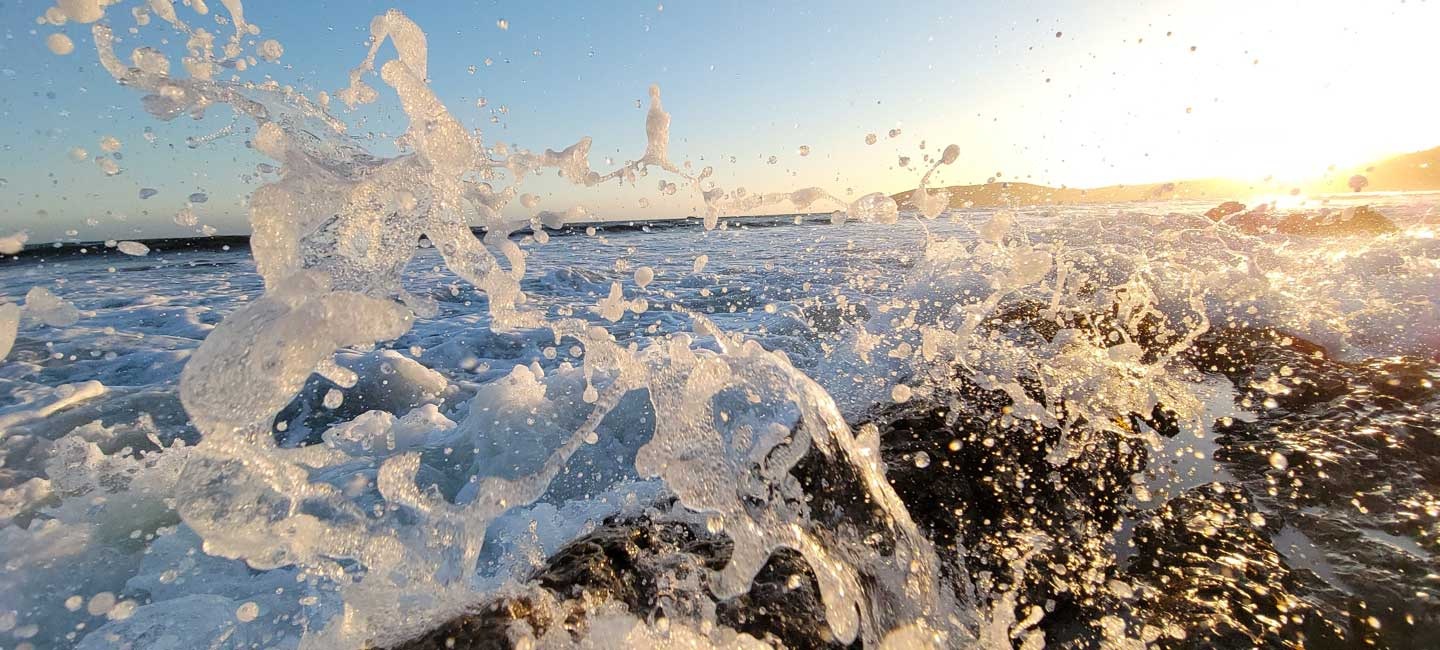 A close up of a wave crashing against rocks in the foreground. Water splashes in front of the camera, lit by the sun hanging low in the sky just off camera.