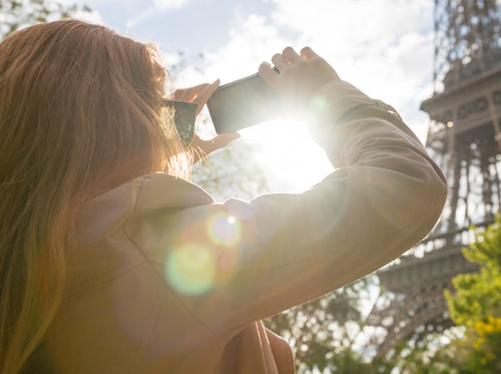 Девушка делает снимки Эйфелевой башни на устройство Galaxy, за башней виден яркий солнечный свет.