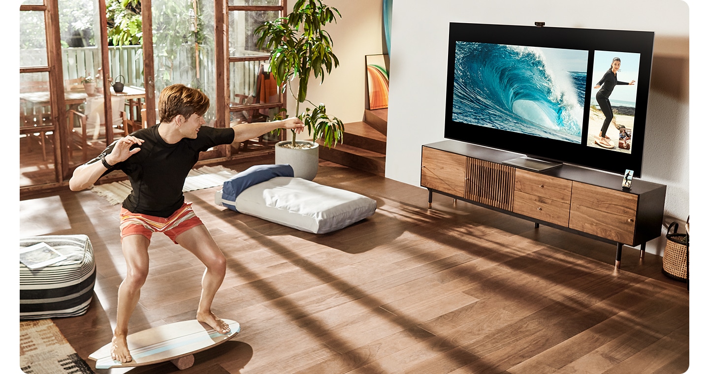 A man is training on a balance board in his living room while receiving real-time instruction from a coach via Multi View on Samsung Smart TV. Using Add View function, he's also looking at ocean waves on the screen for better simulating surfing action.