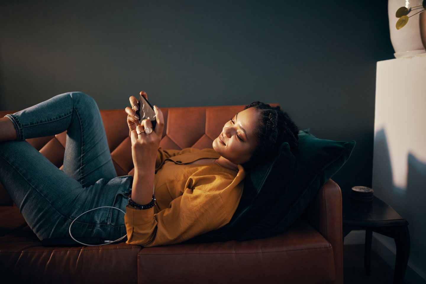 Woman laying on a brown couch in her living room, holding her Galaxy smartphone in landscape mode and she looks at the screen.