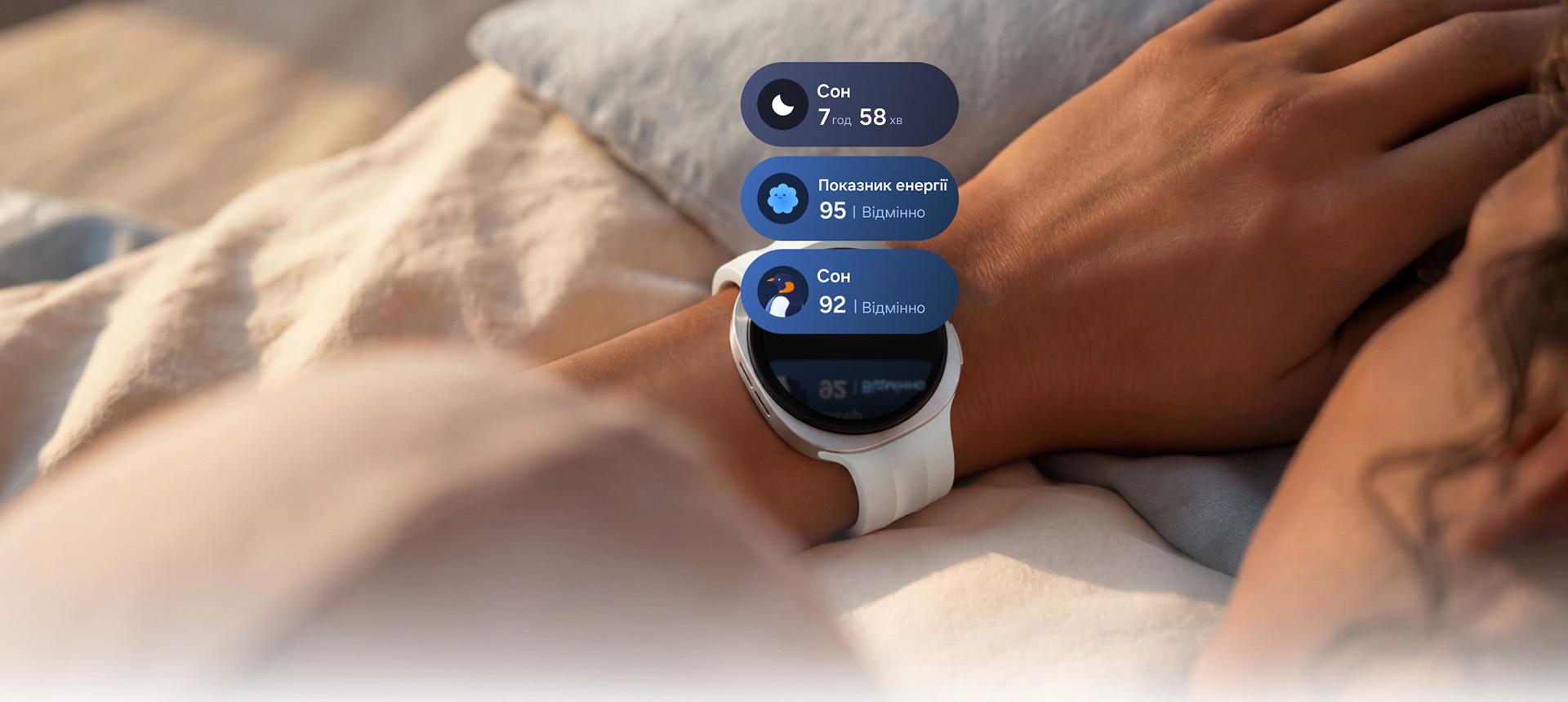 Galaxy Watch9 сріблястого кольору зі Спортивним ремінцем бежевого кольору на зап’ясті людини; від годинника по руці людини розходяться світлові хвилі, що символізують відстеження сну. Над дисплеєм годинника з’являються три картки з даними про сон: картка з тривалістю сну 7 год 58 хв, картка «Показник енергії 92» та картка з порадами щодо сну, на якій зображено піктограму пінгвіна та напис «Перевірте свої звички».