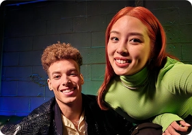 A smiling male wearing a leather jacket and yellow shirt poses next to a red head female with a green top to show off the Galaxy S23 Nightography feature.
