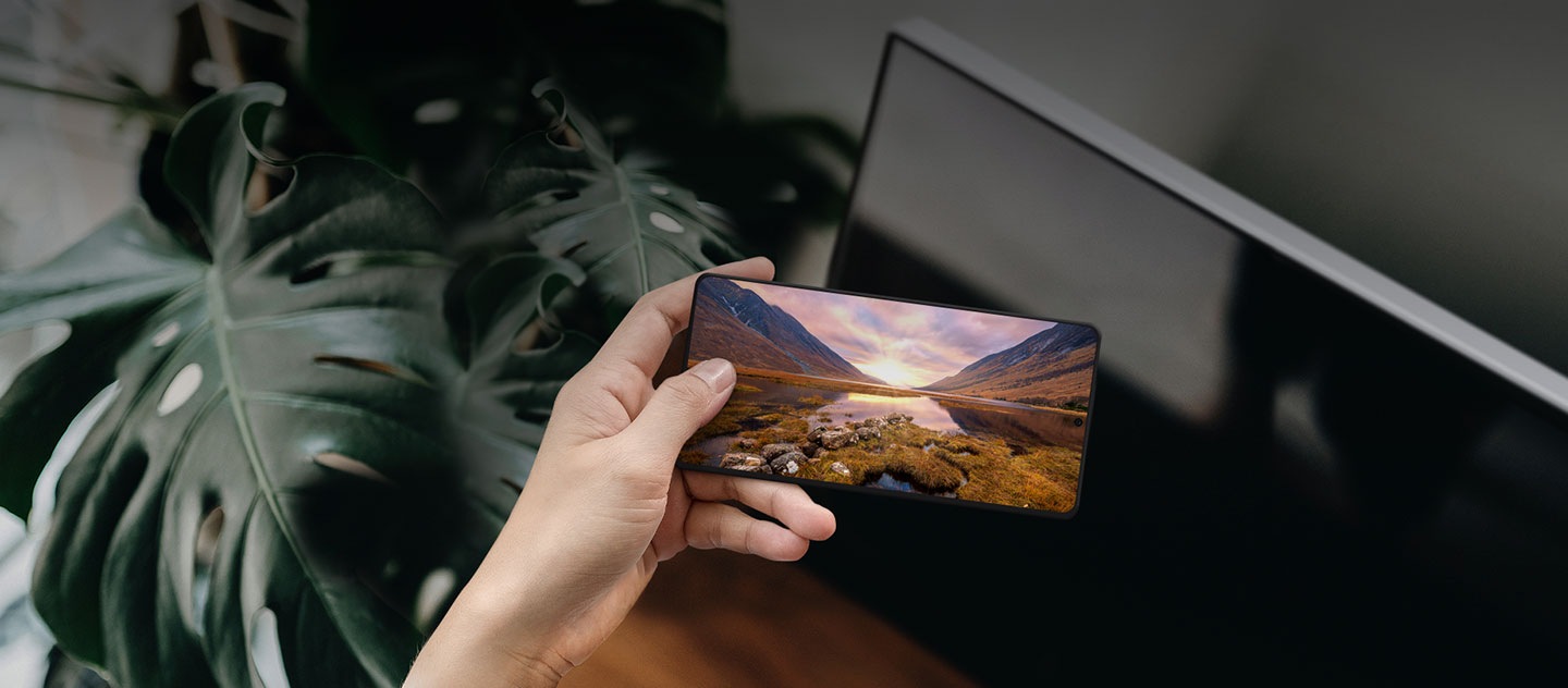 A hand holding a Galaxy smartphone in front of a Samsung TV. The Galaxy screen shows a majestic landscape image.