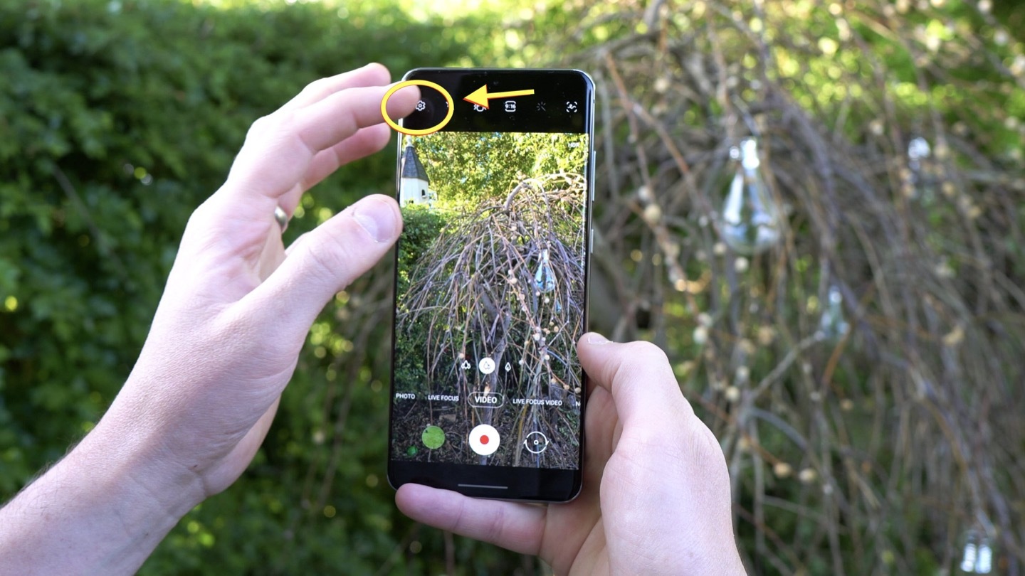 Two hands hold the smartphone Samsung Galaxy S20 Ultra in a portrait orientationin in front of a bush. The camera of the smartphone is on, capturing the bush in front of it. A yellow circle and arrow are pictured around the settings button on the phone screen.