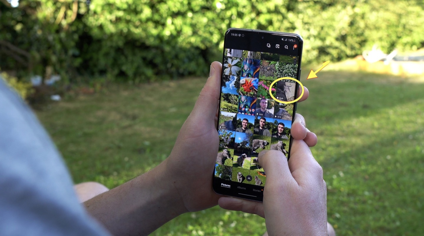 Two hands hold the smartphone Samsung Galaxy S20 Ultra in a portrait orientationin in front of a garden. On the phone screen, a gallery of photos and vifdeos are shown. A yellow circle and arrow are pictured pointing towards one video in the photo and video gallery pictured on screen.