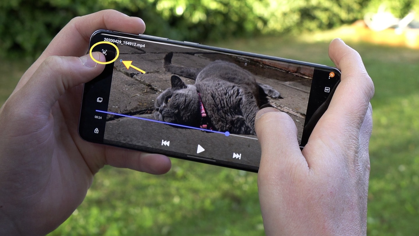 Two hands hold the smartphone Samsung Galaxy S20 Ultra in a landscape orientationin in front of a garden. On the phone screen, a video of a grey cat lying down on the floor is shown, with a yellow circle and arrow arround the photo gallery icon in the top left hand corner of the screen