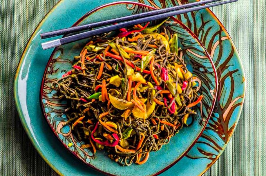 A plate of food with a black noodles, peppers, carrots in a stir fry and a pair of dark blue chopsticks, on a green woven mat