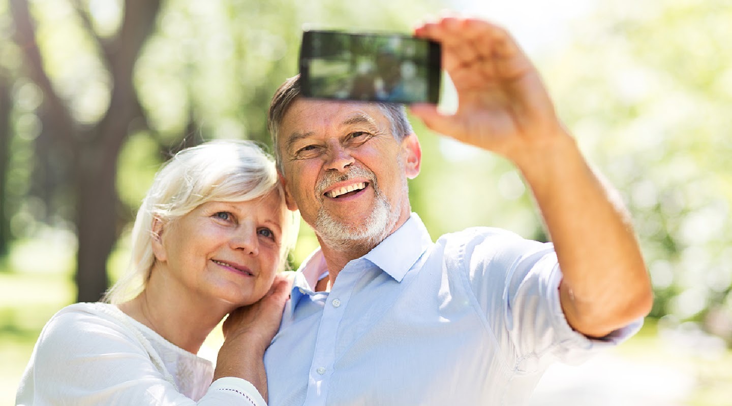 An elderly couple smile while posing for a photo in front of a blurred green background. The man holds a Samsung smartphone in his hand to take the photo.