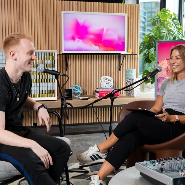 Two live-show hosts are introducing green Galaxy Z Flip3. The female host is holding the product towards her, looking at the cover display. The male host is pointing the side of the product. A camera is live-streaming their demonstration.