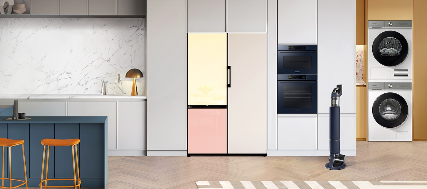 There is a 2 door Bespoke refrigerator with a Vanilla top and Peach bottom, a Beige Bespoke 1 door refrigerator, a built-in oven and a compact oven built into a grey cabinet in a stylish kitchen. A Bespoke Jet in Midnight Blue is standing next to the built-in ovens. There’s a White Bespoke AI™ washer and dryer set built into a yellow wall, with the dryer stacked on top.