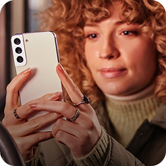 A close up image of a woman with curly ginger hair, holding a Galaxy smartphone to take a picture of the foreground.