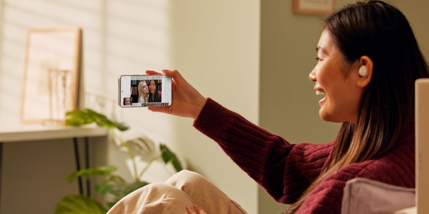 A woman sitting on a bed while on a video call using Samsung Galaxy S22 and Galaxy Buds